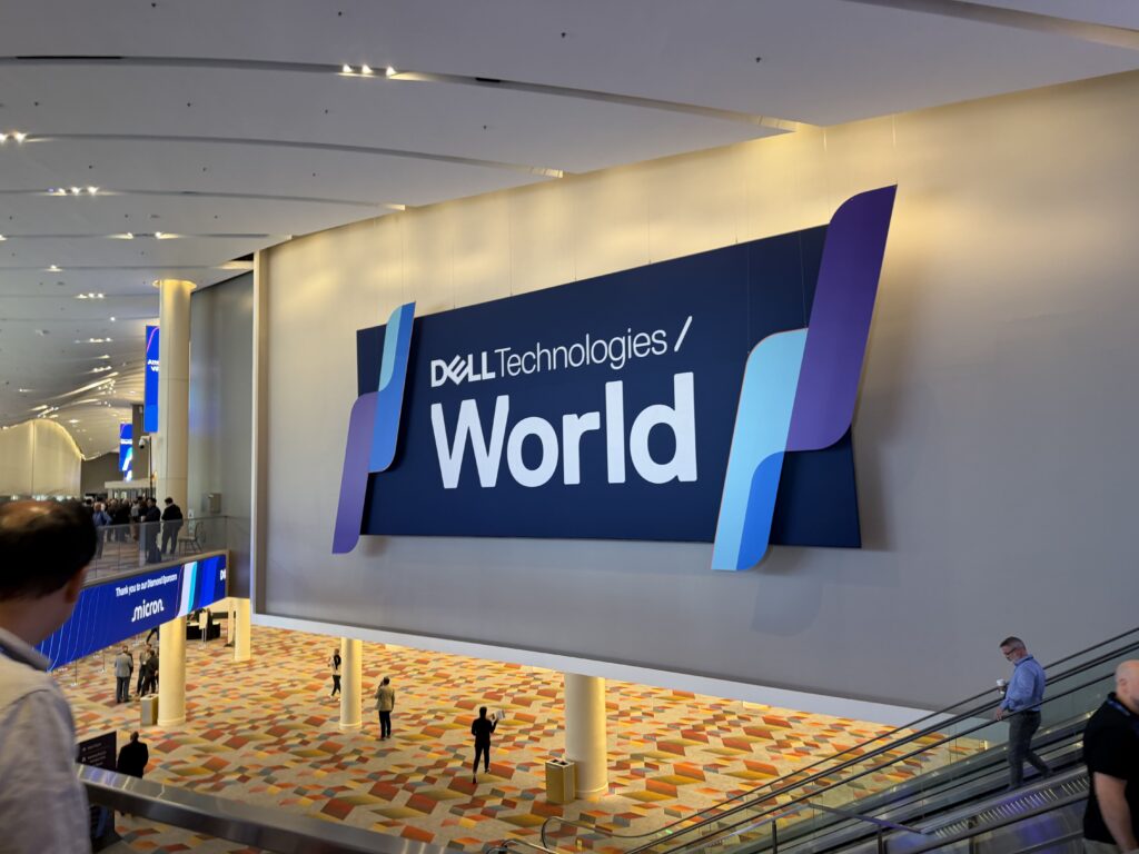 Large ‘Dell Technologies World’ sign displayed in a convention center hall, with attendees walking below and an escalator in the foreground.